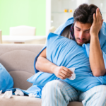 Homem doente coberto com cobertor azul abraçando travesseiro, segurando lenço de papel